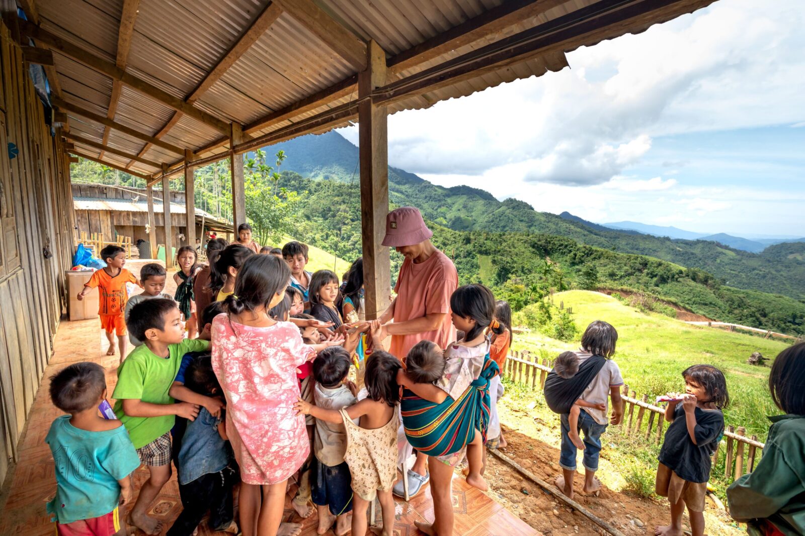 A group of children gather on a terrace overlooking rolling hills and mountains on a sunny day.